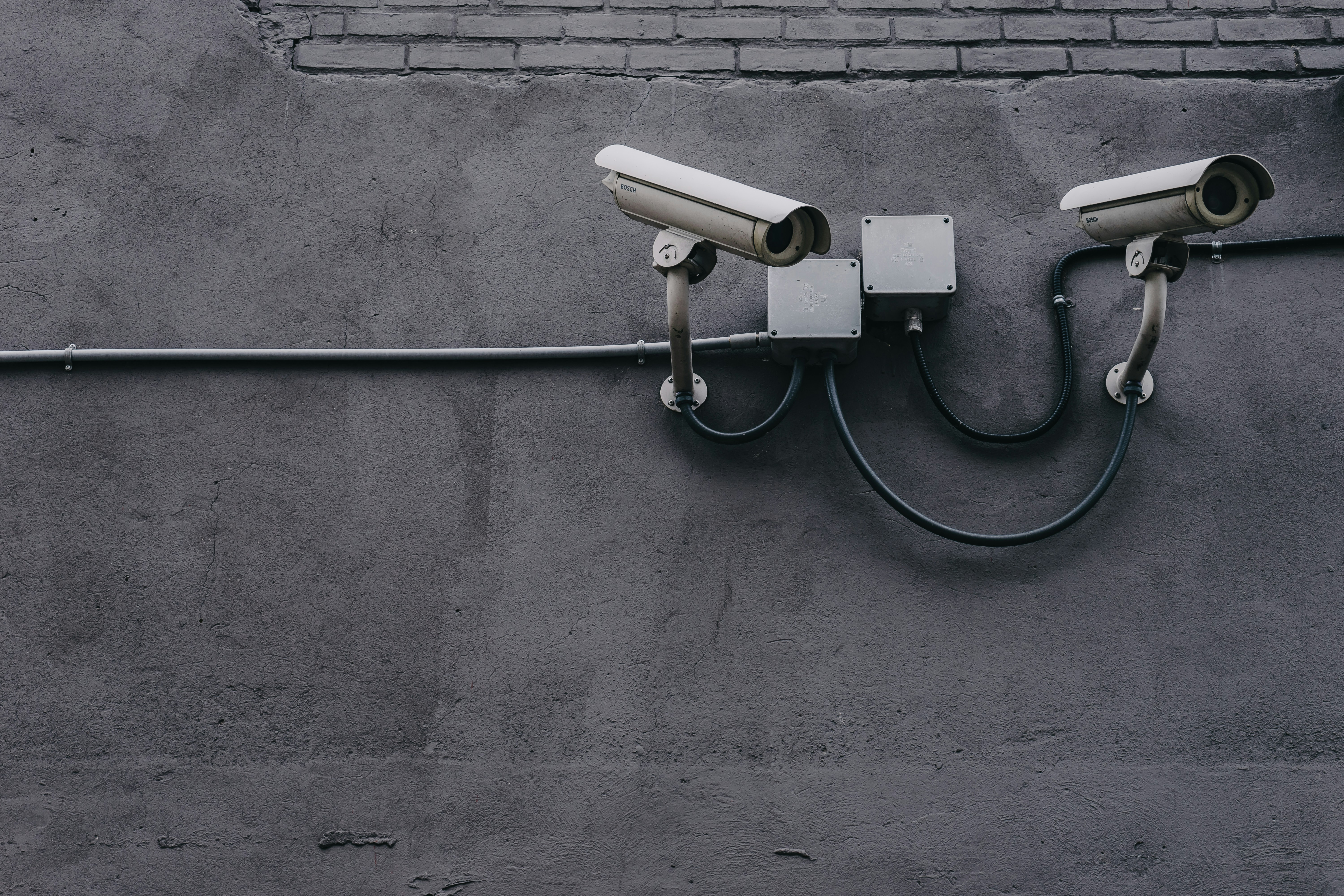 A black and white photograph of two bullet surveillance cameras attached to a concrete and brick wall.